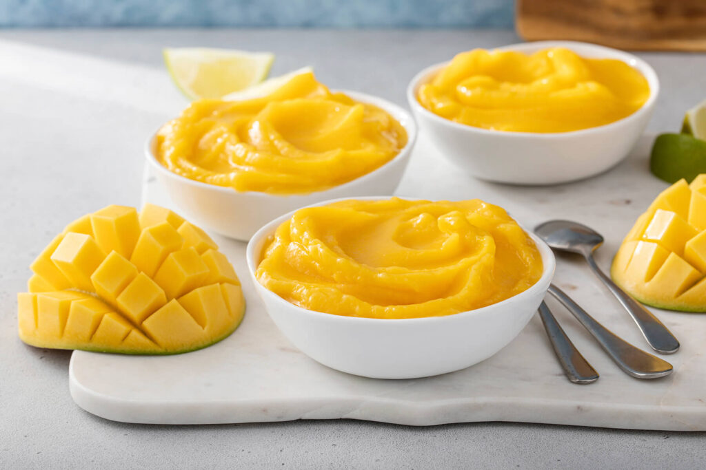 Mehrere weiße Schälchen mit cremiger Mango Nice Cream und eine aufgeschnittene Mango im Igel-Schnitt auf einem Marmorbrett, seitlich fotografiert