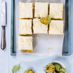 Blick von oben auf ein blaugraues Holztablett mit Backpapier und sieben geschnittenen Mango-Schnitten: hell elfenbeinfarbene Oberfläche mit dunklen Vanillepünktchen und flach angedrückten groben Spänen weißer Schokolade, darunter die schmale gelbe Mangoschicht und der helle Boden, daneben ein altes Messer sowie Physalis in einer weißen Schale auf hellblauem Holz