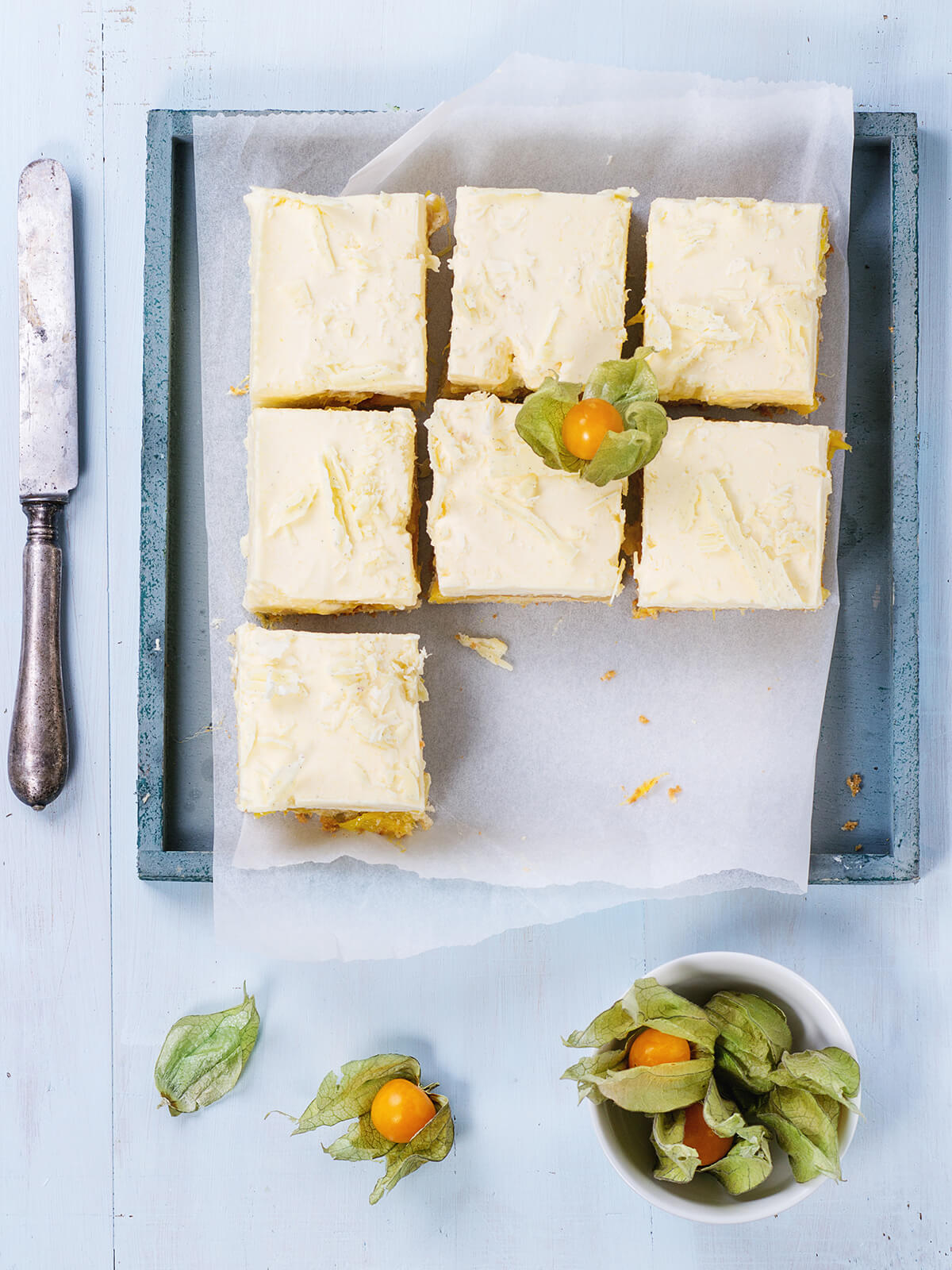 Blick von oben auf ein blaugraues Holztablett mit Backpapier und sieben geschnittenen Mango-Schnitten: hell elfenbeinfarbene Oberfläche mit dunklen Vanillepünktchen und flach angedrückten groben Spänen weißer Schokolade, darunter die schmale gelbe Mangoschicht und der helle Boden, daneben ein altes Messer sowie Physalis in einer weißen Schale auf hellblauem Holz