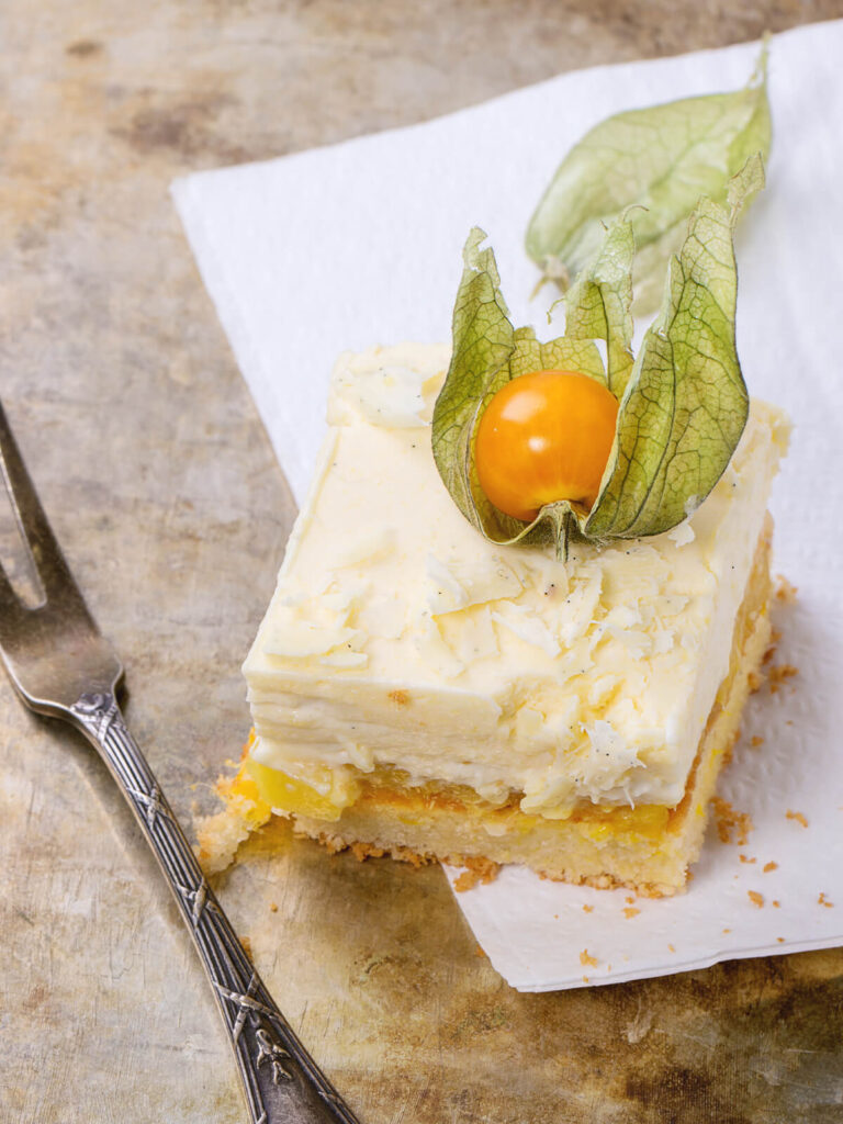 Einzelne Mango-Schnitte auf einer weißen Serviette auf hellem Steinuntergrund: heller goldgelber Boden, gelbe Mangoschicht mit sichtbaren Fruchtstücken und dicke hell elfenbeinfarbene Schokoladencreme mit dunklen Vanillepünktchen und groben Spänen weißer Schokolade, der Anschnitt der Creme dicht und in feinen Lagen, obenauf eine Physalis mit geöffneten grünen Kelchblättern, links eine silberne Kuchengabel