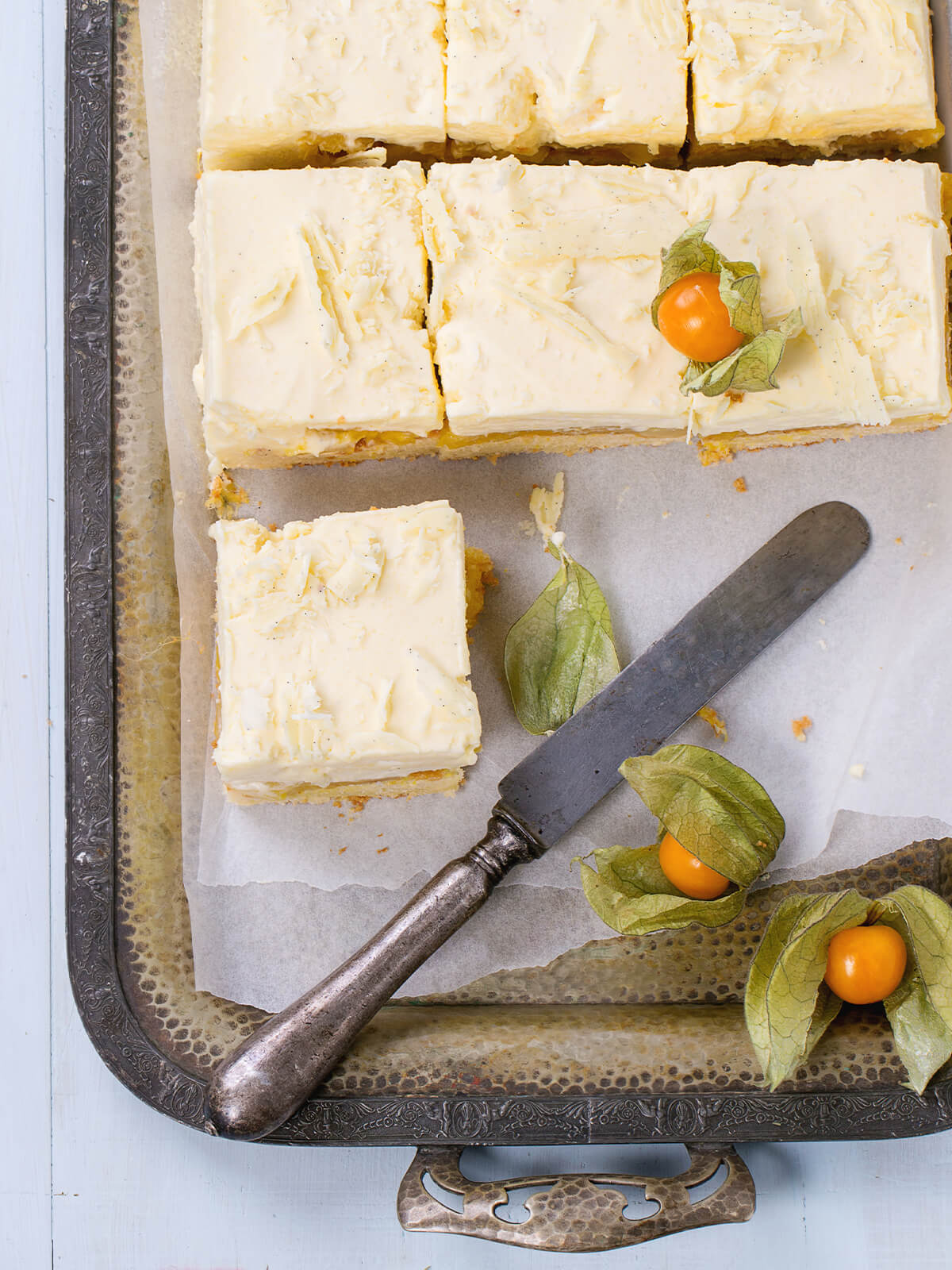 Blick von oben auf ein verziertes silbernes Tablett mit Backpapier und mehreren geschnittenen Mango-Schnitten mit hell elfenbeinfarbener Oberfläche, dunklen Vanillepünktchen und flach eingedrückten groben Spänen weißer Schokolade, ein Stück abgerückt, daneben ein antikes Messer und mehrere Physalis auf hellblauem Holz
