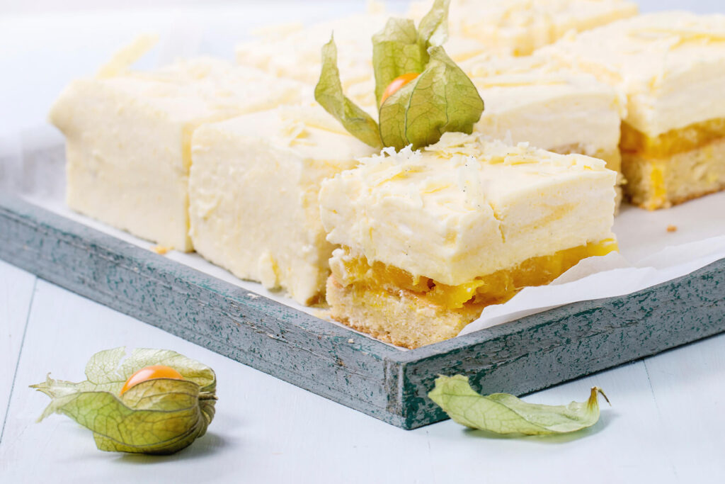 Fertige Mango-Schnitten auf Backpapier auf einem graugrünen Holztablett, ein Stück nach vorn gestellt: heller goldgelber Boden, glänzend gelbe Mangoschicht mit sichtbaren Fruchtstücken und dicke hell elfenbeinfarbene Schokoladencreme mit dunklen Vanillepünktchen und groben Spänen weißer Schokolade, der Anschnitt der Creme dicht und in feinen Lagen, davor Physalis mit grünen Kelchblättern auf weiß gestrichenem Holz