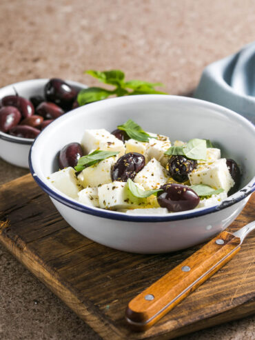 Marinierter Feta in Würfeln mit Kalamata-Oliven, Basilikum und Oregano in einer Emailleschale, daneben eine Schale Oliven