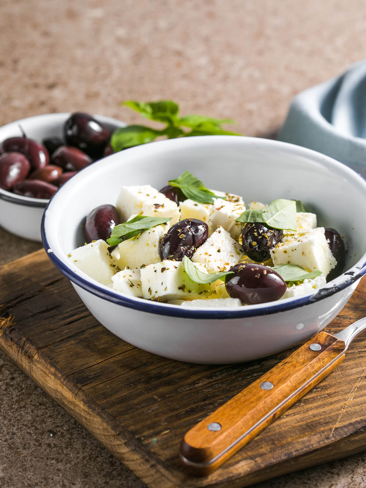 Marinierter Feta in Würfeln mit Kalamata-Oliven, Basilikum und Oregano in einer Emailleschale, daneben eine Schale Oliven