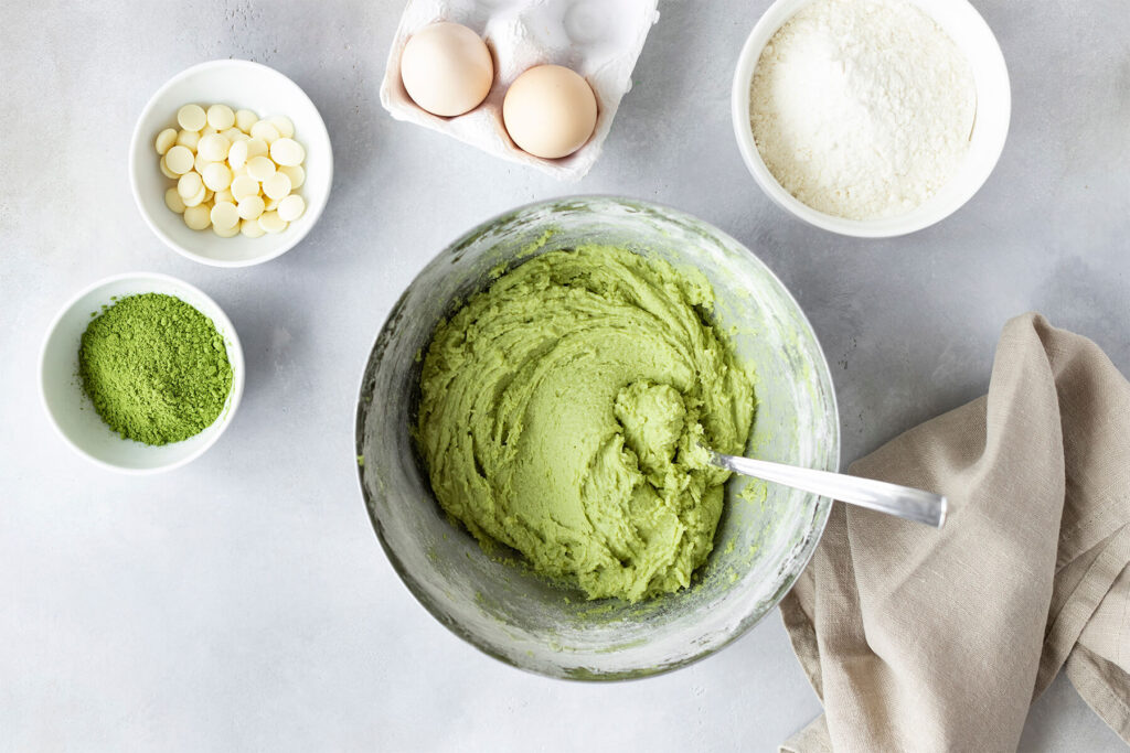 Grüner Matcha-Keksteig in einer Rührschüssel, daneben Matcha-Pulver, Eier, Mehl und weiße Schokotropfen