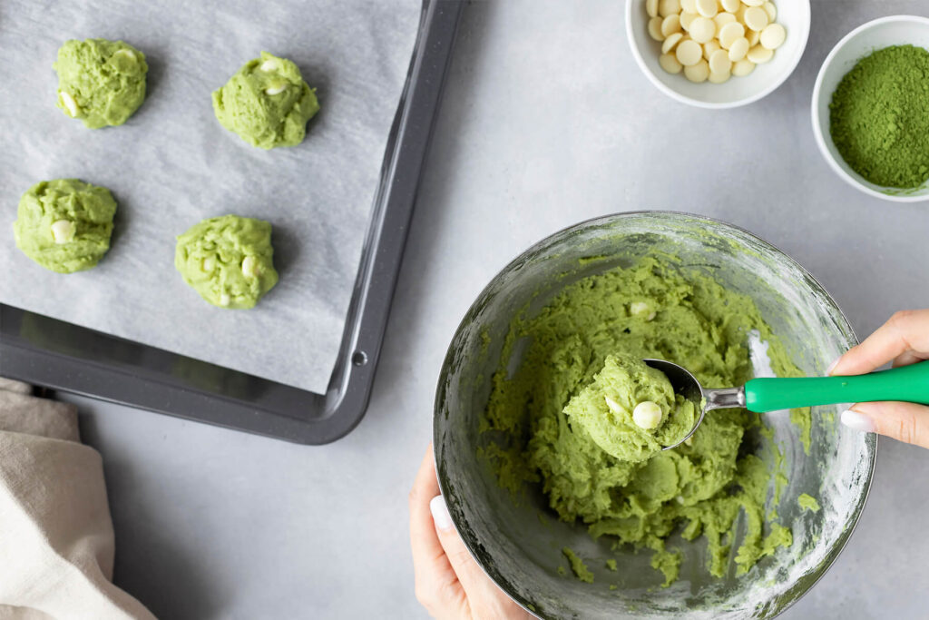 Grüner Matcha-Keksteig wird mit einem Portionierer aus der Schüssel abgestochen, im Hintergrund ein Blech mit Teighäufchen