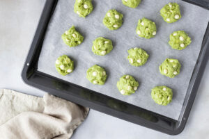 Rohe grüne Matcha-Teighäufchen mit weißen Schokotropfen mit Abstand auf einem Backblech