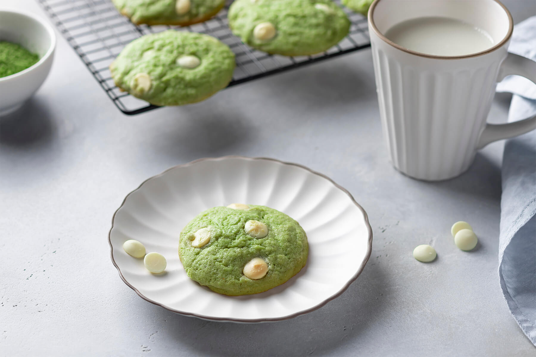 Ein einzelner grüner Matcha-Cookie auf einem Teller, daneben ein Becher Milch und weitere Cookies auf einem Kühlgitter