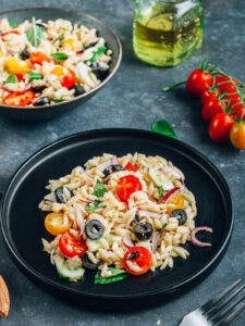 Mediterraner Orzo-Salat auf einem schwarzen Teller mit glänzenden Orzo-Nudeln, halbierten roten und gelben Kirschtomaten, Gurke, roten Zwiebeln, schwarzen Olivenringen und frischem Basilikum