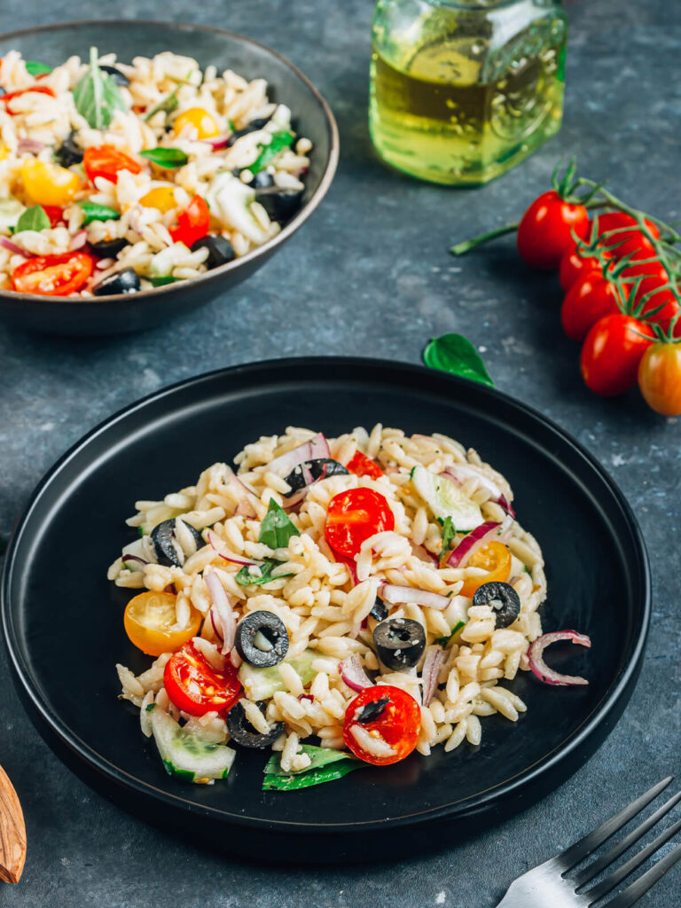 Mediterraner Orzo-Salat auf einem schwarzen Teller mit glänzenden Orzo-Nudeln, halbierten roten und gelben Kirschtomaten, Gurke, roten Zwiebeln, schwarzen Olivenringen und frischem Basilikum
