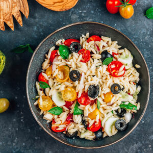Mediterraner Orzo-Salat von oben in einer dunklen Schüssel mit Orzo-Nudeln, halbierten roten und gelben Kirschtomaten, Gurke, roten Zwiebeln, schwarzen Olivenringen und frischem Basilikum in glänzendem Olivenöl-Dressing