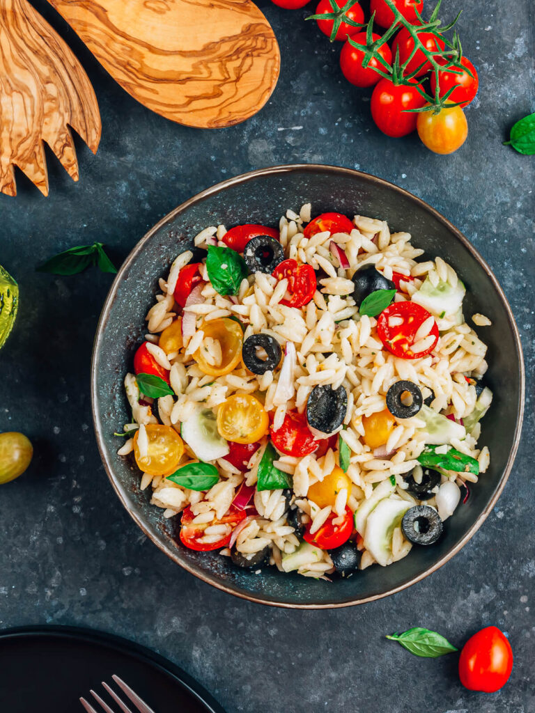 Mediterraner Orzo-Salat von oben in einer dunklen Schüssel mit Orzo-Nudeln, halbierten roten und gelben Kirschtomaten, Gurke, roten Zwiebeln, schwarzen Olivenringen und frischem Basilikum in glänzendem Olivenöl-Dressing
