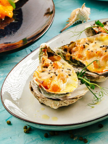 Überbackene Meeresfrüchte-Julienne mit goldgelbem Käse in mehreren Austernschalen; die vordere ist mit schwarzem Sesam bestreut, daneben liegt ein Zweig Erbsensprossen