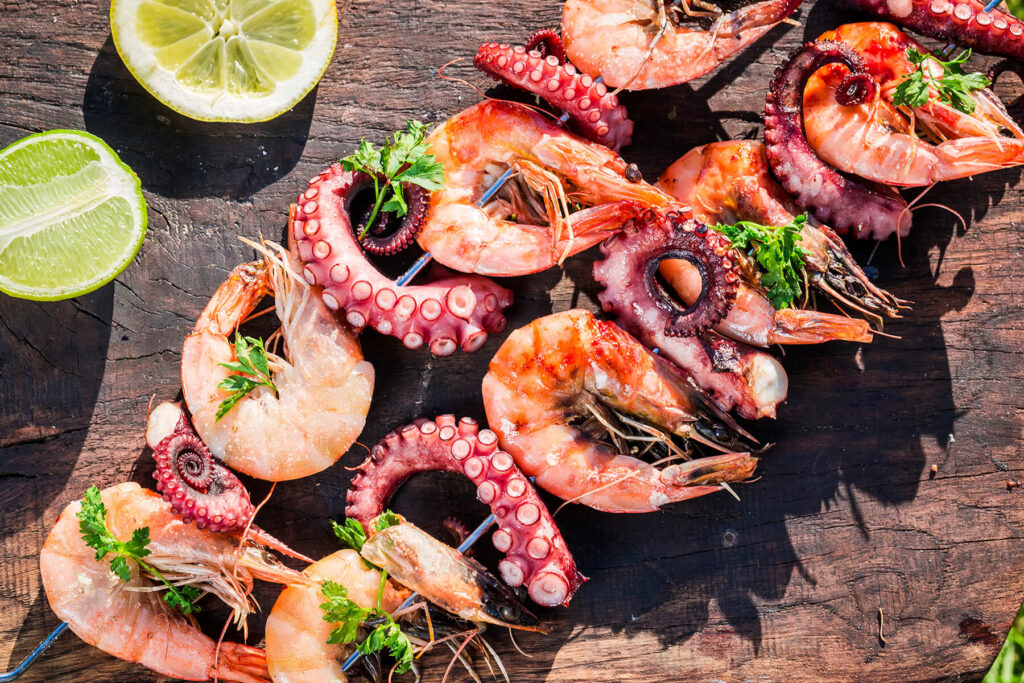 Gegrillte Meeresfrüchte-Spieße mit Garnelen und Oktopus, Petersilie und Zitrone auf einem Holzbrett