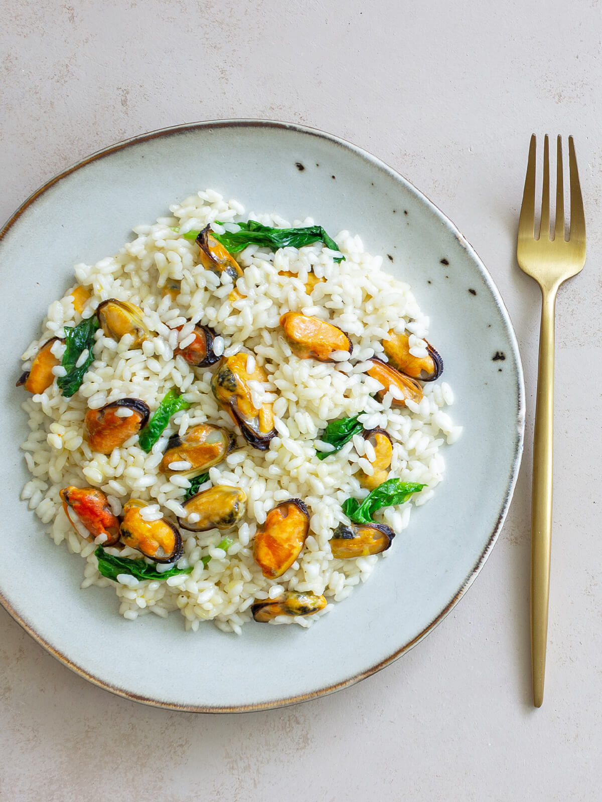 Cremiges Risotto mit ausgelösten Miesmuscheln und frischem Spinat auf einem blaugrauen Teller von oben, dazu eine goldene Gabel