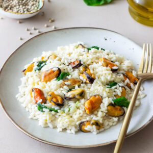 Cremiges Risotto mit ausgelösten Miesmuscheln und frischem Spinat auf einem blaugrauen Teller, dazu eine goldene Gabel