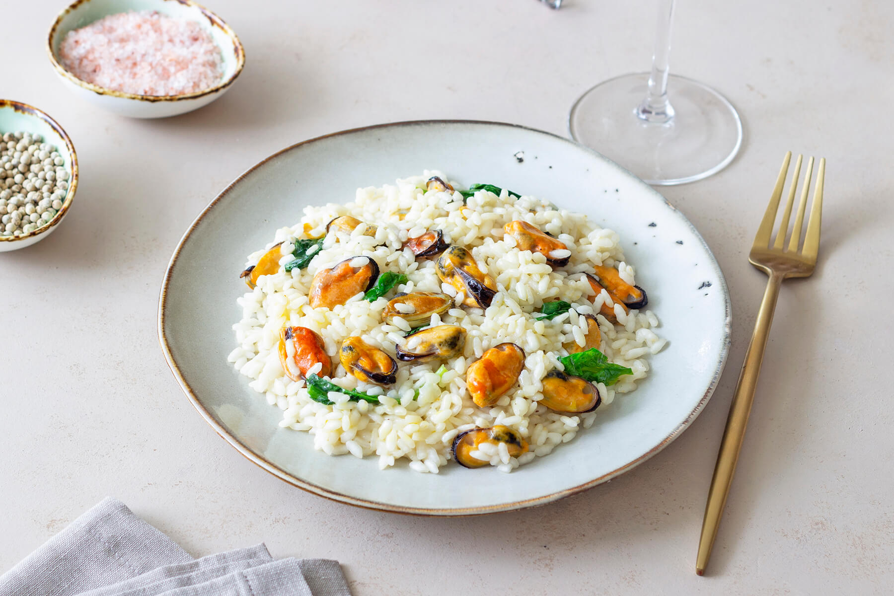 Cremiges Miesmuschel-Risotto mit Spinat auf einem Teller, daneben Salz, weißer Pfeffer und ein Weinglas