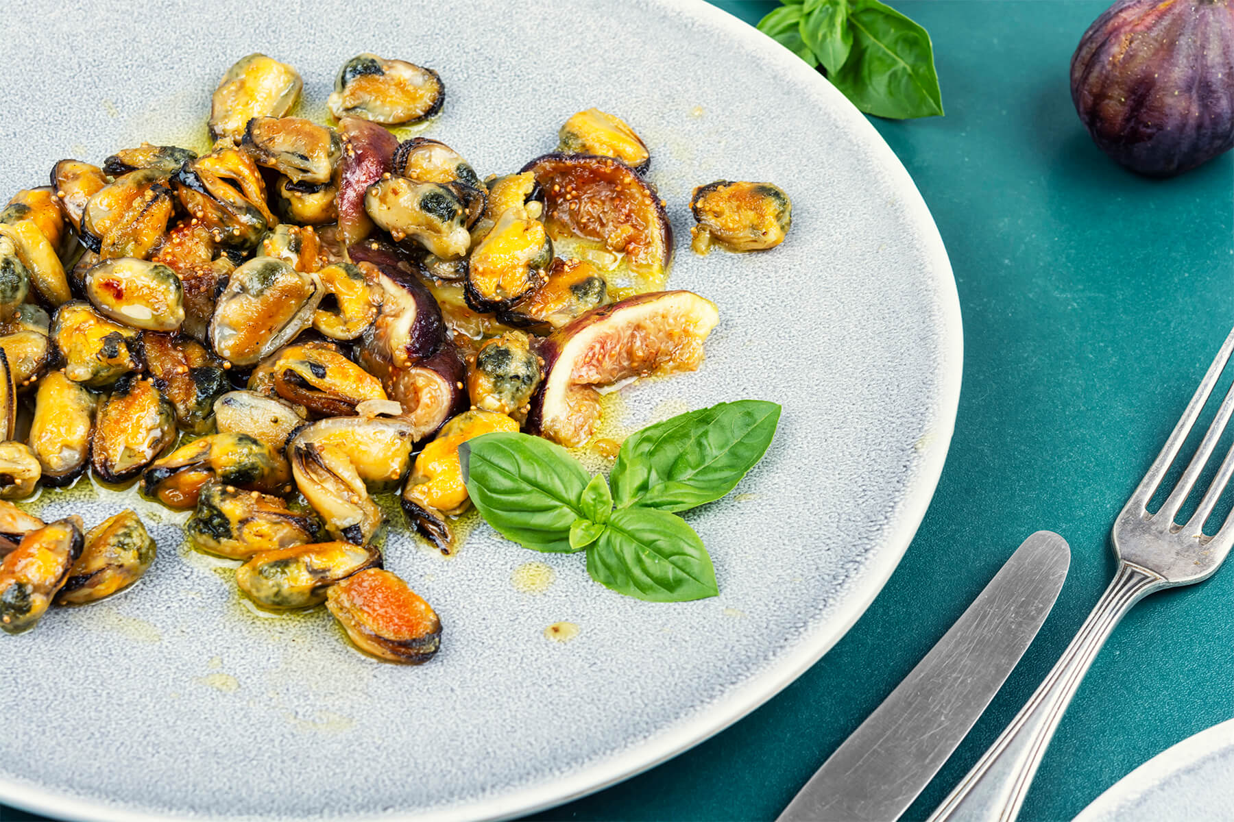 Nahaufnahme eines hellblau-grauen, gesprenkelten Steinguttellers mit gebratenem Muschelfleisch in Goldorange, dunklen Röstkanten und dunklen Mantelrändern, dazwischen Feigenspalten mit violetter Schale und rosa Fruchtfleisch mit sichtbaren feinen Kernen, glänzender goldener Ölsud und frische grüne Basilikumblätter; rechts petrolfarbener Untergrund mit altem Messer, alter Gabel und einer ganzen dunklen Feige