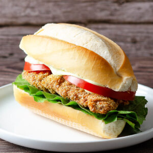 Knusprige argentinische Rinder-Milanesa mit Salat und Tomate in einem länglichen Brötchen auf weißem Teller vor Holzhintergrund