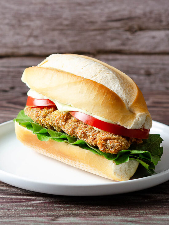 Knusprige argentinische Rinder-Milanesa mit Salat und Tomate in einem länglichen Brötchen auf weißem Teller vor Holzhintergrund