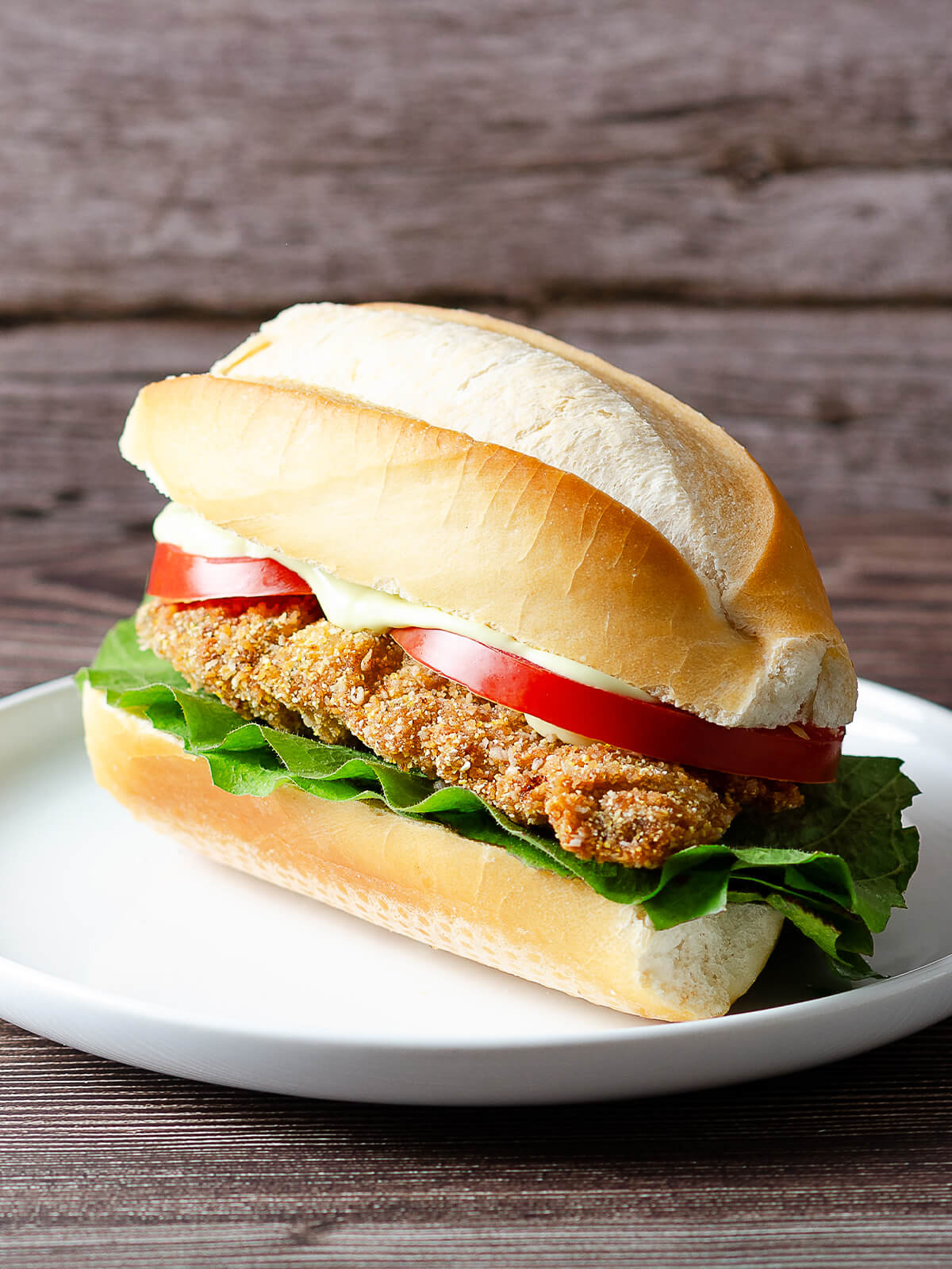 Knusprige argentinische Rinder-Milanesa mit Salat und Tomate in einem länglichen Brötchen auf weißem Teller vor Holzhintergrund