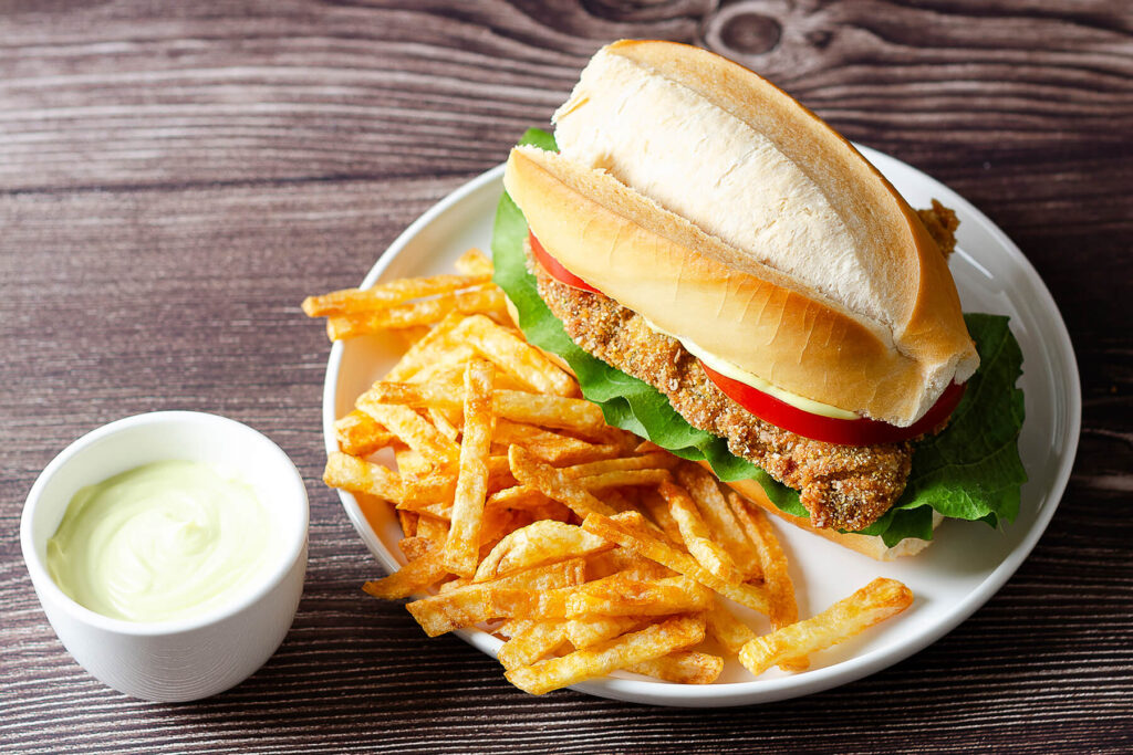 Argentinisches Milanesa-Sandwich mit Salat und Tomate im Brötchen, dazu knusprige Pommes frites und ein Schälchen Mayonnaise-Dip