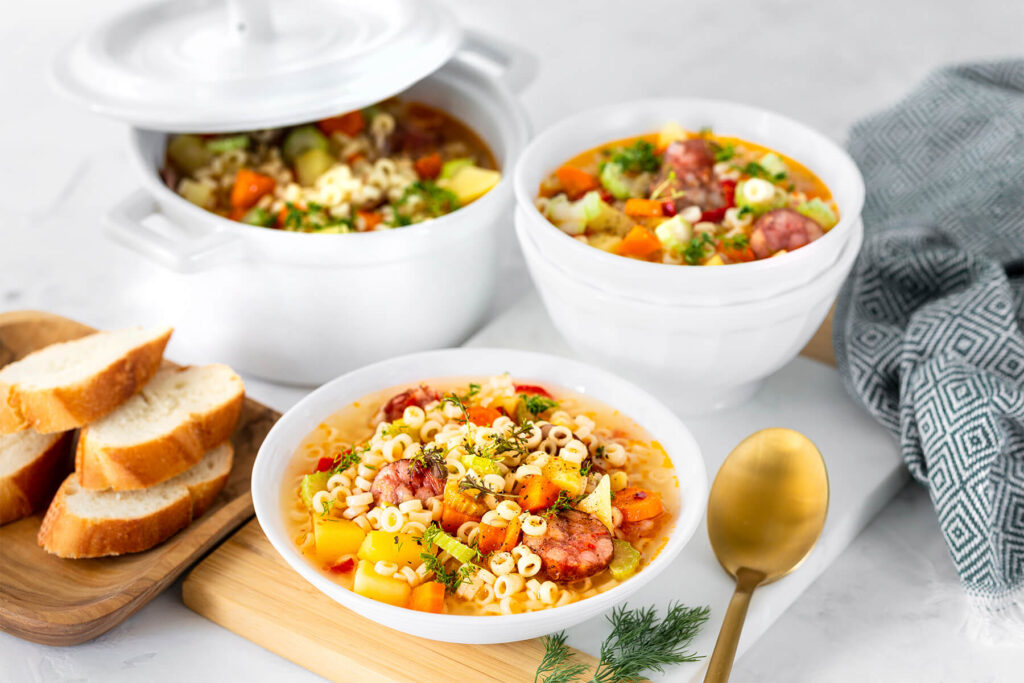 Weißer tiefer Teller mit klarer goldgelber Suppe auf einem Holzbrett, darin helle Nudelringe, blassgelbe Kartoffelwürfel, orange Karottenscheiben, hellgrüne Selleriestücke und Scheiben geräucherter Wurst mit gebräuntem Rand, obenauf feine Kräuter und grob gemahlener Pfeffer; dahinter ein weißer Topf mit angelehntem Deckel voller Suppe und eine weiße Schale, links Weißbrotscheiben auf einem Holzbrett, rechts ein goldfarbener Löffel und ein grau-weiß gemustertes Tuch, vorn ein Dillzweig