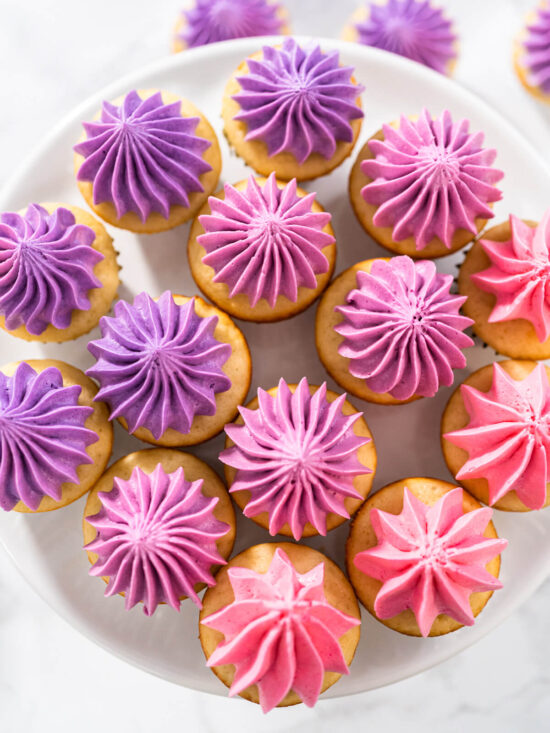 Mini-Vanille-Cupcakes mit Buttercreme im Ombre-Verlauf von Violett über Beere bis Rosa auf einer weißen Kuchenplatte