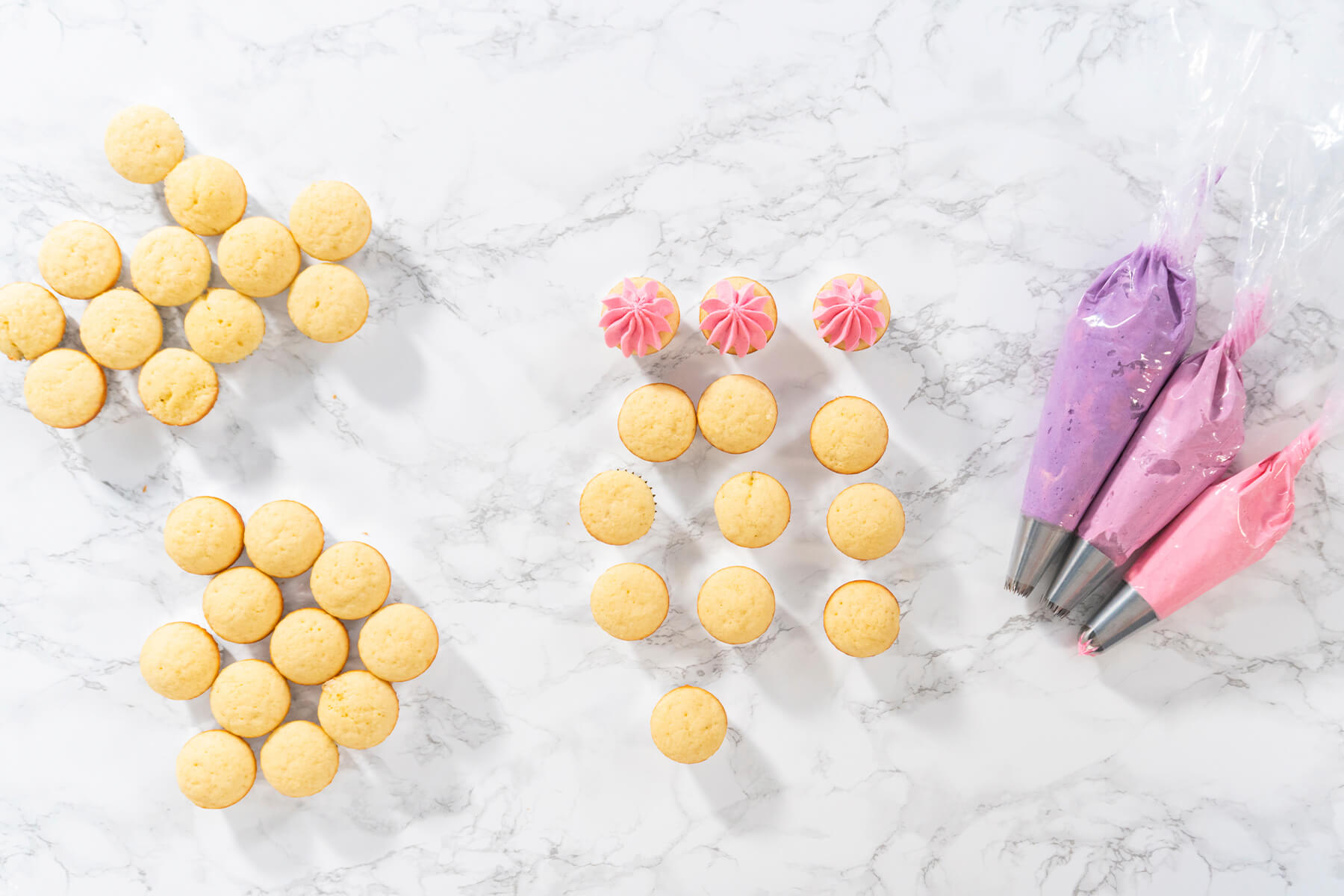 Von oben unverzierte Mini-Cupcakes, rosa gespritzte Cupcakes und drei Spritzbeutel in Violett, Beere und Rosa