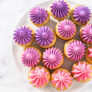 Viele Mini-Vanille-Cupcakes mit Buttercreme im Farbverlauf von Violett über Beere bis Rosa auf einer weißen Kuchenplatte von oben