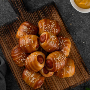 Mini-Würstchen im Hefeteig auf einem Holzbrett von oben, kleine Würstchenstücke spiralförmig in goldbraunen Hefeteig gewickelt und mit Sesam bestreut, die runden Würstchen-Schnittflächen schauen an den offenen Enden heraus, dazu Schälchen mit Senf und Ketchup