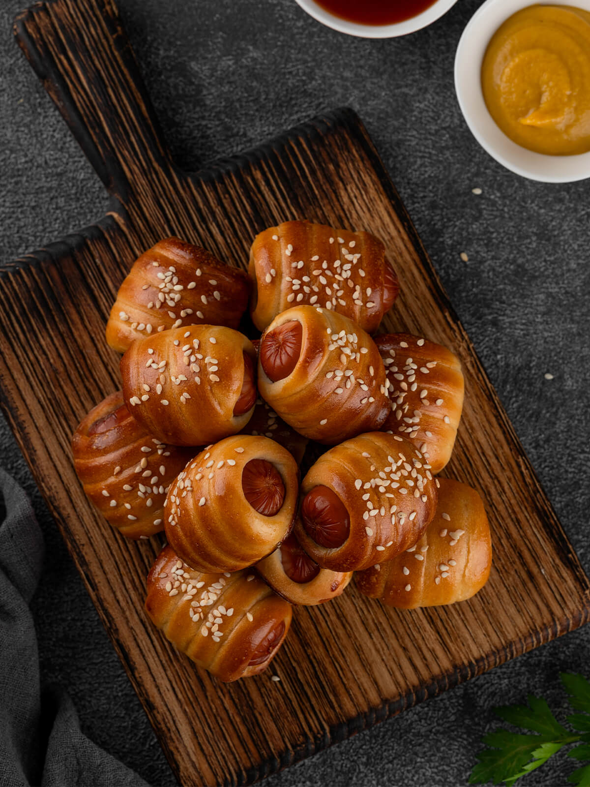 Mini-Würstchen im Hefeteig auf einem Holzbrett von oben, kleine Würstchenstücke spiralförmig in goldbraunen Hefeteig gewickelt und mit Sesam bestreut, die runden Würstchen-Schnittflächen schauen an den offenen Enden heraus, dazu Schälchen mit Senf und Ketchup