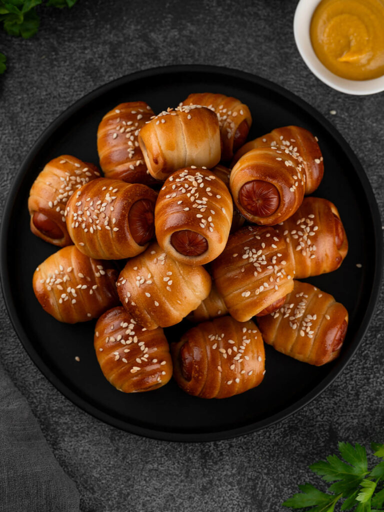 Ein Teller mit Mini-Würstchen im Hefeteig hochkant von oben, kleine Würstchenstücke spiralförmig in goldbraunen, glänzenden Hefeteig gewickelt und mit Sesam bestreut, die runden Würstchen-Schnittflächen schauen an den offenen Enden heraus, dazu eine Schale Senf