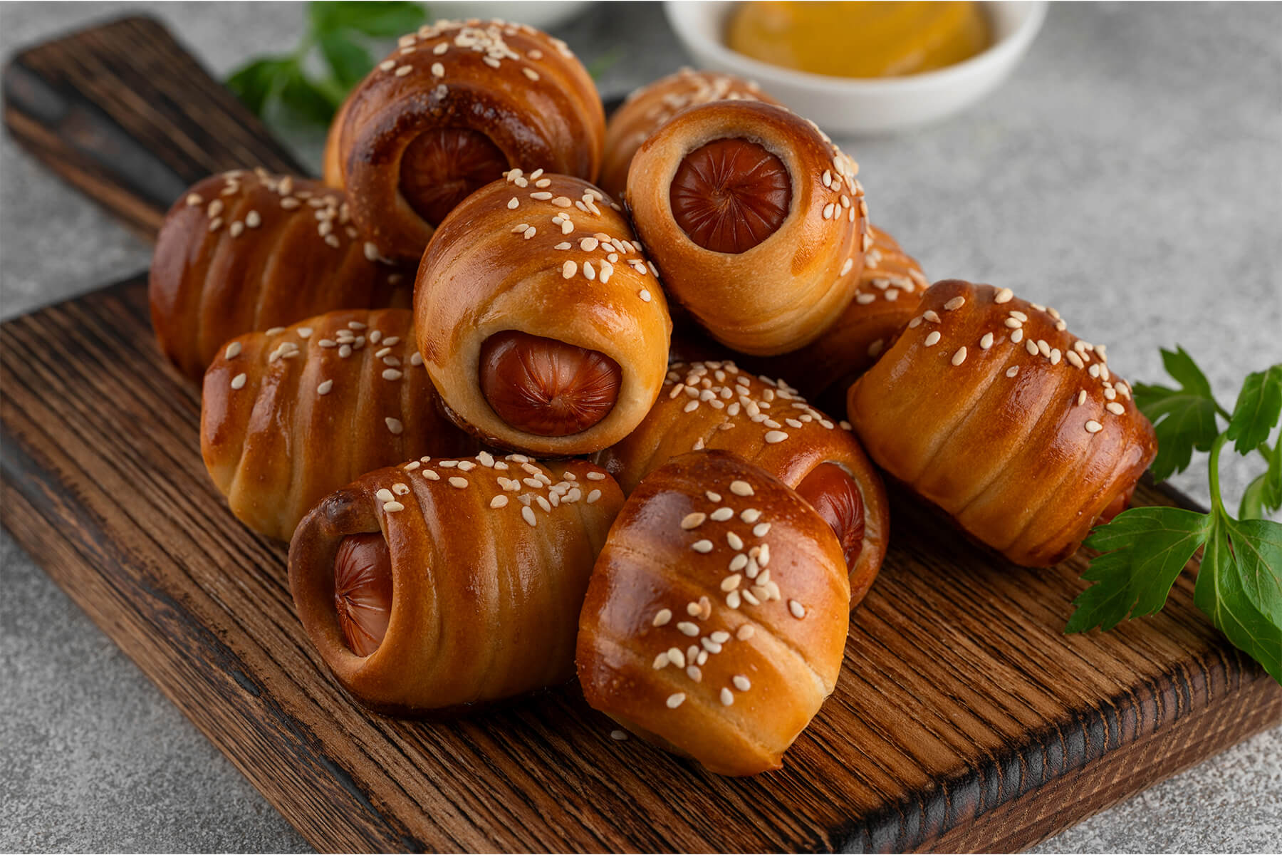 Mini-Würstchen im Hefeteig auf einem Holzbrett in Nahaufnahme, spiralförmig umwickelte Würstchenstücke mit Sesam bestreut, die runden Würstchen-Schnittflächen an den offenen Enden sichtbar, dazu ein Schälchen Senf