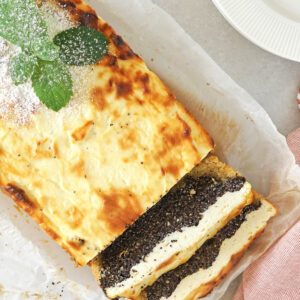 Angeschnittener Mohn-Quark-Kuchen als Kastenkuchen mit goldener Kruste, daneben zwei Scheiben mit dünnem Mürbeteigboden, dicker dunkler Mohnschicht und heller Quarkmasse darüber, bestäubt mit Puderzucker und mit Minze garniert