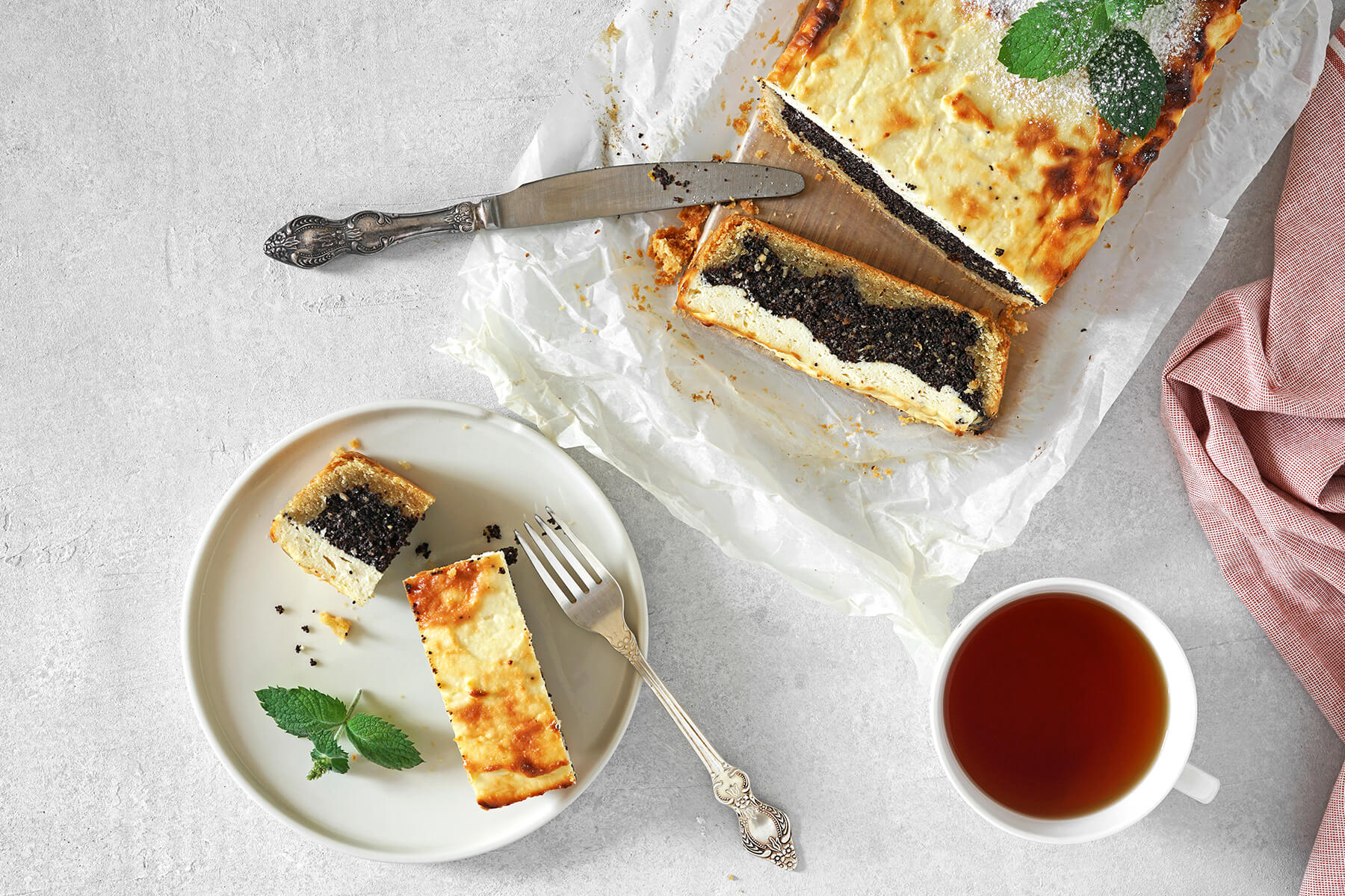 Ganzer Mohn-Quark-Kuchen als Laib auf Backpapier mit Messer und einer abgeschnittenen Scheibe, daneben ein Teller mit zwei Scheiben und eine Tasse Tee, die goldene Kruste sowie dünner Mürbeteigboden, dicke dunkle Mohnschicht und helle Quarkmasse darüber gut erkennbar