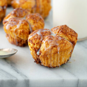 Mehrere Zimt-Zucker Monkey-Bread-Muffins auf einer Marmorplatte, oben aus vier bis fünf goldbraunen, dick mit Zimt-Zucker bestreuten Kuppeln zusammengesetzt, an den Flanken die feinen senkrechten Rillen des abgezogenen Papierförmchens, mit dünnem weißem Zuckerguss beträufelt, daneben eine Flasche Milch und Zimtstangen