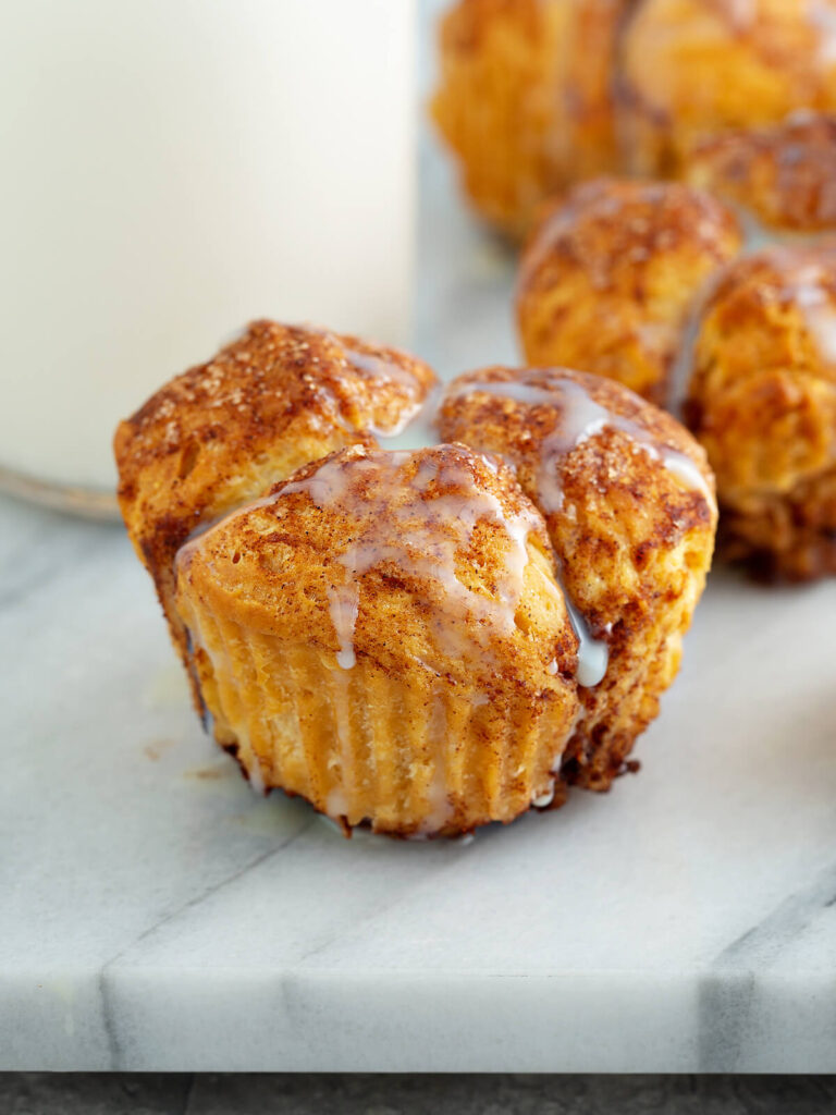 Ein einzelner Zimt-Zucker Monkey-Bread-Muffin in Nahaufnahme auf einer Marmorplatte, aus mehreren goldbraunen, in Zimt-Zucker gewälzten Teigstücken mit sichtbaren blättrigen Schichten zusammengesetzt und mit weißem Zuckerguss beträufelt