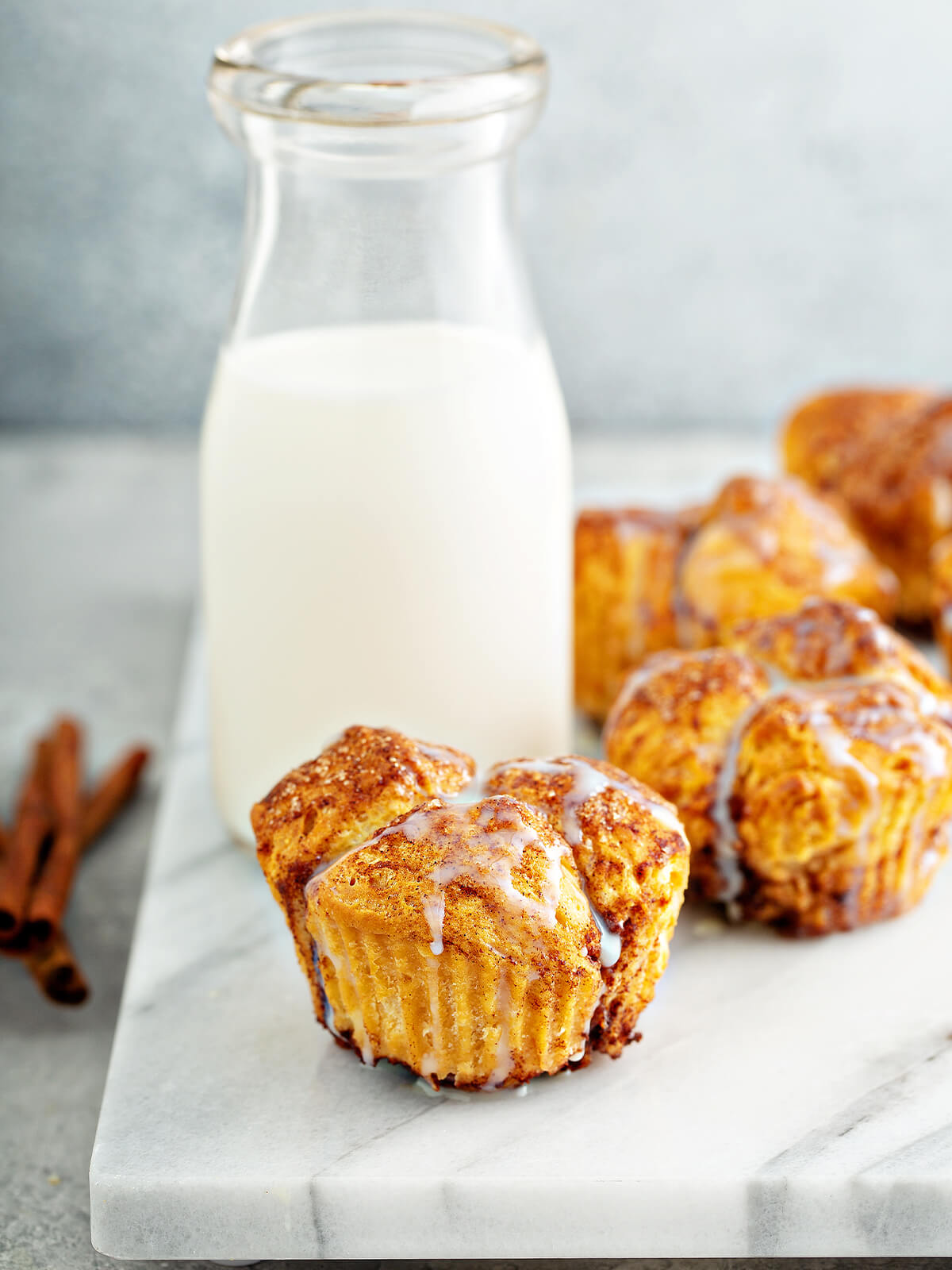 Zimt-Zucker Monkey-Bread-Muffins auf einer hellen Marmorplatte, oben aus vier bis fünf goldbraunen, dick mit Zimt-Zucker bestreuten Kuppeln zusammengesetzt, an den Flanken die feinen senkrechten Rillen des abgezogenen Papierförmchens, mit dünnem weißem Zuckerguss in schmalen Rinnsalen beträufelt, dahinter eine Flasche Milch, im Vordergrund links ein paar Zimtstangen