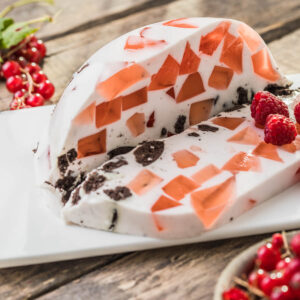 Angeschnittene Kuppel einer Beeren-Mosaik-Torte auf weißer rechteckiger Platte, schräg von oben fotografiert: in weißer Sauerrahm-Masse sitzen unregelmäßige durchscheinende Scherben aus Beerengelee in Koralle und Apricot, dazwischen dunkle Keksbrösel, das abgeschnittene Stück liegt vorn, rechts drei Himbeeren, links oben eine Rispe roter Johannisbeeren