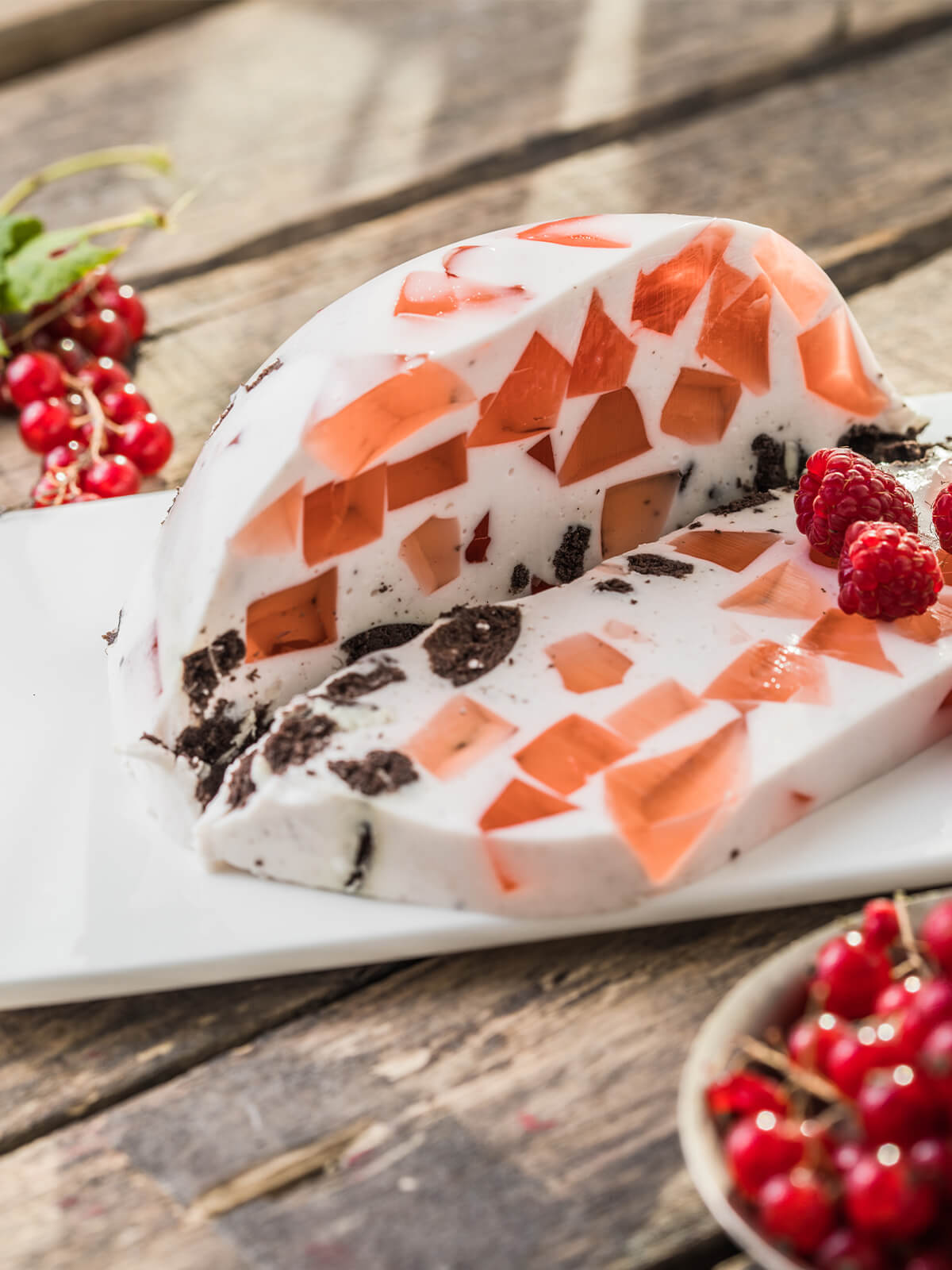 Angeschnittene Kuppel einer Beeren-Mosaik-Torte auf weißer rechteckiger Platte, schräg von oben fotografiert: in weißer Sauerrahm-Masse sitzen unregelmäßige durchscheinende Scherben aus Beerengelee in Koralle und Apricot, dazwischen dunkle Keksbrösel, das abgeschnittene Stück liegt vorn, rechts drei Himbeeren, links oben eine Rispe roter Johannisbeeren
