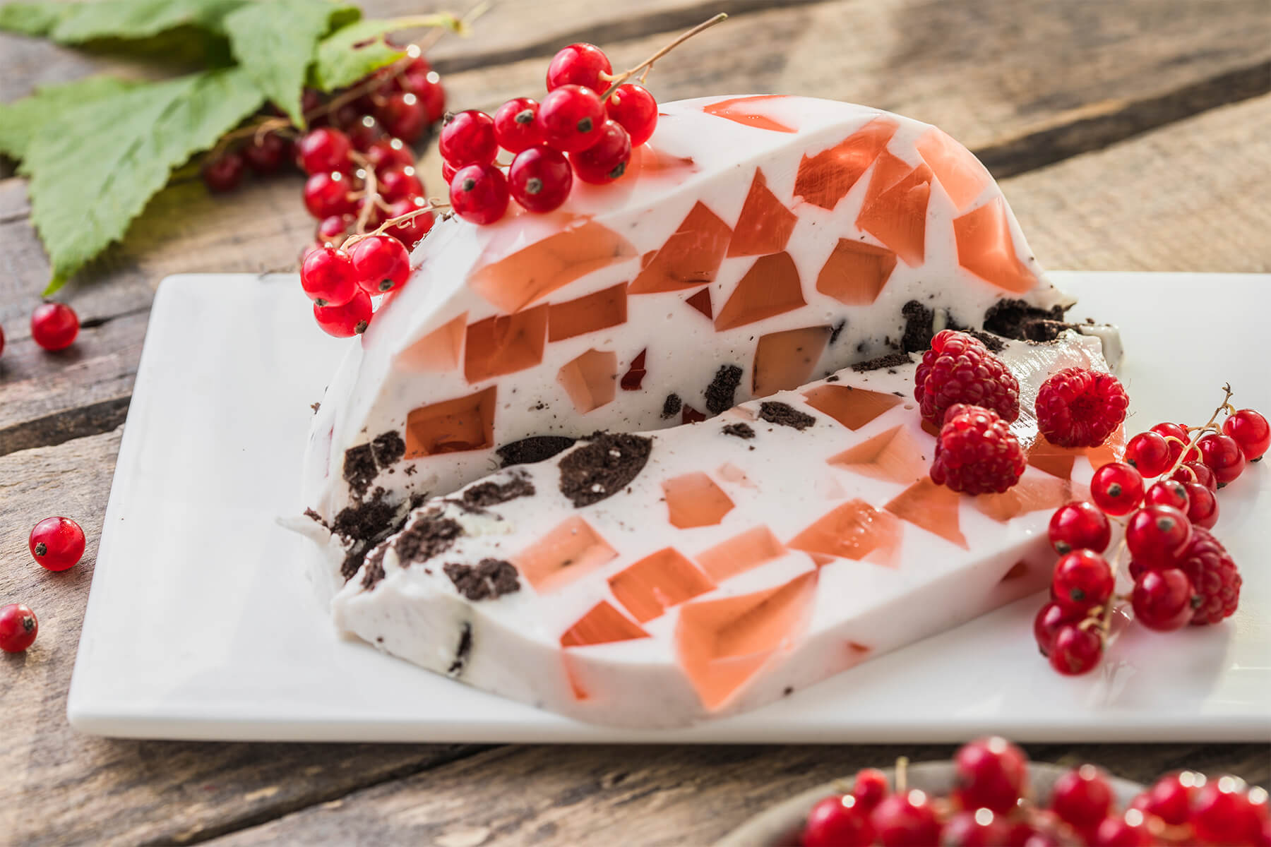 Beeren-Mosaik-Torte im Anschnitt von der Seite auf weißer rechteckiger Platte: unregelmäßige, durchscheinende Scherben aus Beerengelee in Koralle und Apricot sitzen in weißer Sauerrahm-Masse, entlang der Unterseite eine unregelmäßige Schicht dunkler Schokokeks-Brösel, darüber Rispen roter Johannisbeeren und drei Himbeeren