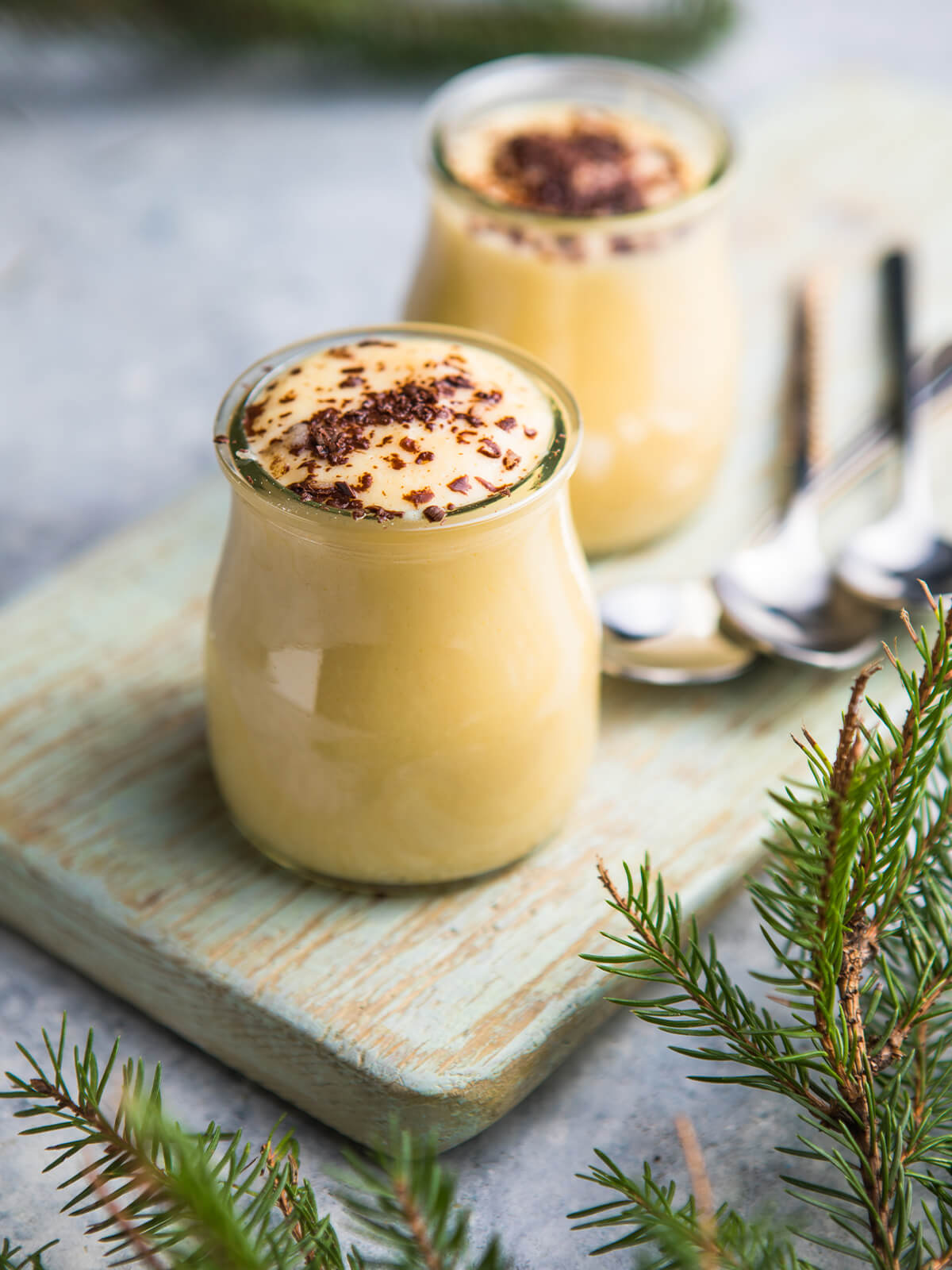Zwei kleine bauchige Gläser mit spanischem Milchpudding auf einem hellgrün gestrichenen Holzbrett: die Creme steht randvoll und leicht gewölbt über dem Glasrand, die Oberfläche blassgelb und glatt, die Masse hinter dem Glas goldgelb; in der Mitte liegen locker gestreute mittelgrobe Späne dunkler Schokolade, die braune Schlieren in die feuchte Oberfläche ziehen, der äußere Ring bleibt frei; rechts dahinter zwei silberne Löffel, ringsum Tannenzweige auf hellem Steinuntergrund