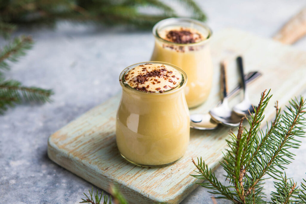 Zwei kleine bauchige Gläser spanischer Milchpudding auf einem hellgrün gestrichenen Holzbrett: die Creme steht randvoll und leicht gewölbt über dem Glasrand, die Oberfläche blassgelb und glatt, die Masse hinter dem Glas goldgelb; obenauf locker in die Mitte gestreute mittelgrobe Späne dunkler Schokolade mit braunen Schlieren ringsherum, der äußere Rand bleibt frei, daneben zwei silberne Löffel mit dunklen Griffen, ringsum Tannenzweige auf hellgrauem Steinuntergrund