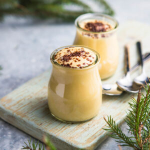 Zwei kleine bauchige Gläser spanischer Milchpudding auf einem hellgrün gestrichenen Holzbrett: die Creme steht randvoll und leicht gewölbt über dem Glasrand, die Oberfläche blassgelb und glatt, die Masse hinter dem Glas goldgelb; obenauf locker in die Mitte gestreute mittelgrobe Späne dunkler Schokolade mit braunen Schlieren ringsherum, der äußere Rand bleibt frei, daneben zwei silberne Löffel mit dunklen Griffen, ringsum Tannenzweige auf hellgrauem Steinuntergrund
