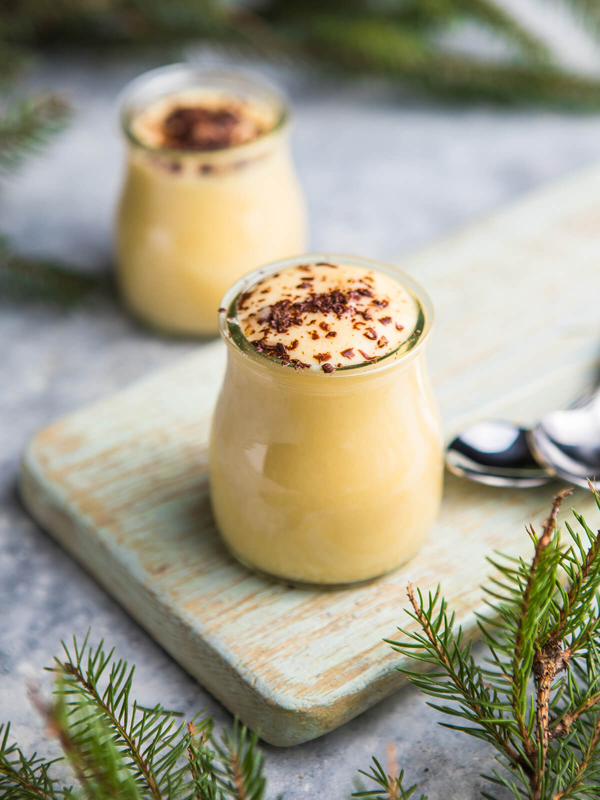 Zwei kleine bauchige Gläser mit spanischem Milchpudding, vorn eines scharf im Bild: die blassgelbe Oberfläche steht glatt, porenfrei und als flache Kuppe leicht über dem Glasrand, die Masse hinter dem Glas wirkt goldgelb; in der Mitte ein locker gehäuftes Nest mittelgrober Schokoladenspäne mit einzelnen Locken, drumherum vereinzelte Splitter und braune Schlieren, der äußere Ring der Creme bleibt frei; das vordere Glas steht auf einem hellgrün gestrichenen Holzbrett, rechts daneben liegen zwei silberne Löffel, das zweite Glas und die Tannenzweige dahinter stehen auf hellgrauem Steinuntergrund