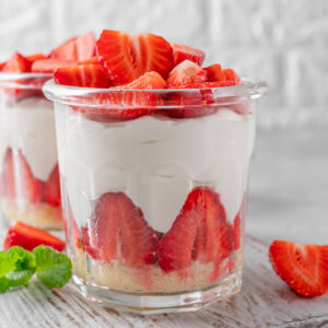 No-Bake Erdbeer-Cheesecake in zwei großen Gläsern mit hellem Keksboden, einem Kranz halbierter Erdbeeren an der Glaswand, cremiger Frischkäsecreme und einem Berg frisch halbierter Erdbeeren obenauf