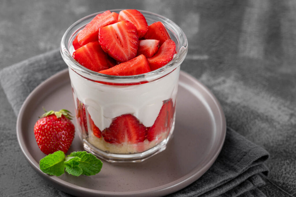 Ein Glas No-Bake Erdbeer-Cheesecake auf einem Teller mit hellem Keksboden, sichtbarem Erdbeer-Kranz, cremiger Frischkäsecreme und frisch geviertelten Erdbeeren obenauf, dazu Minze