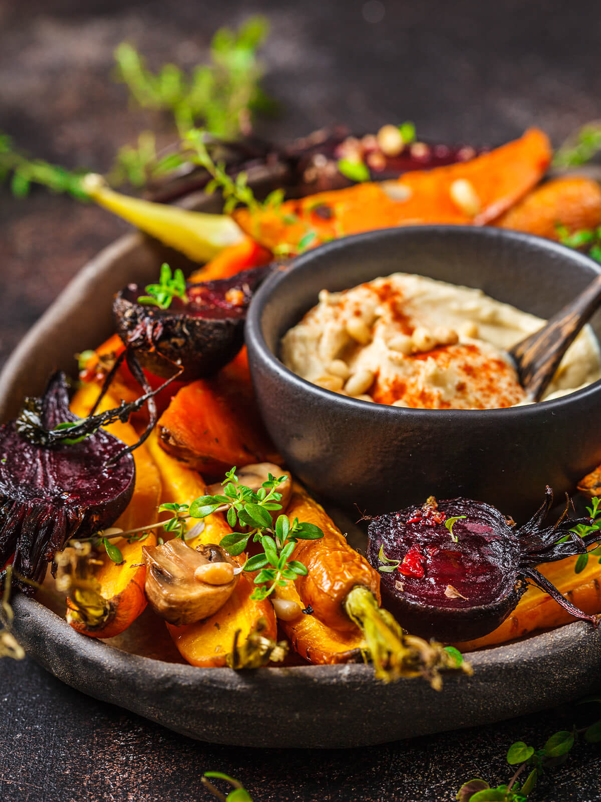 Buntes Ofengemüse mit halbierter roter Bete, Karotten und Champignons auf einem dunklen Teller, daneben eine Schale Hummus
