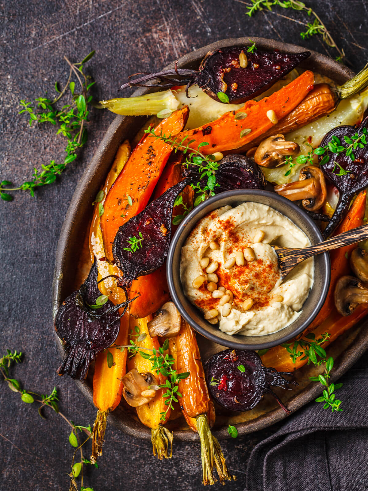 Buntes Ofengemüse von oben auf einem dunklen Teller angerichtet, mit Thymian, Pinienkernen und einer Schale Hummus